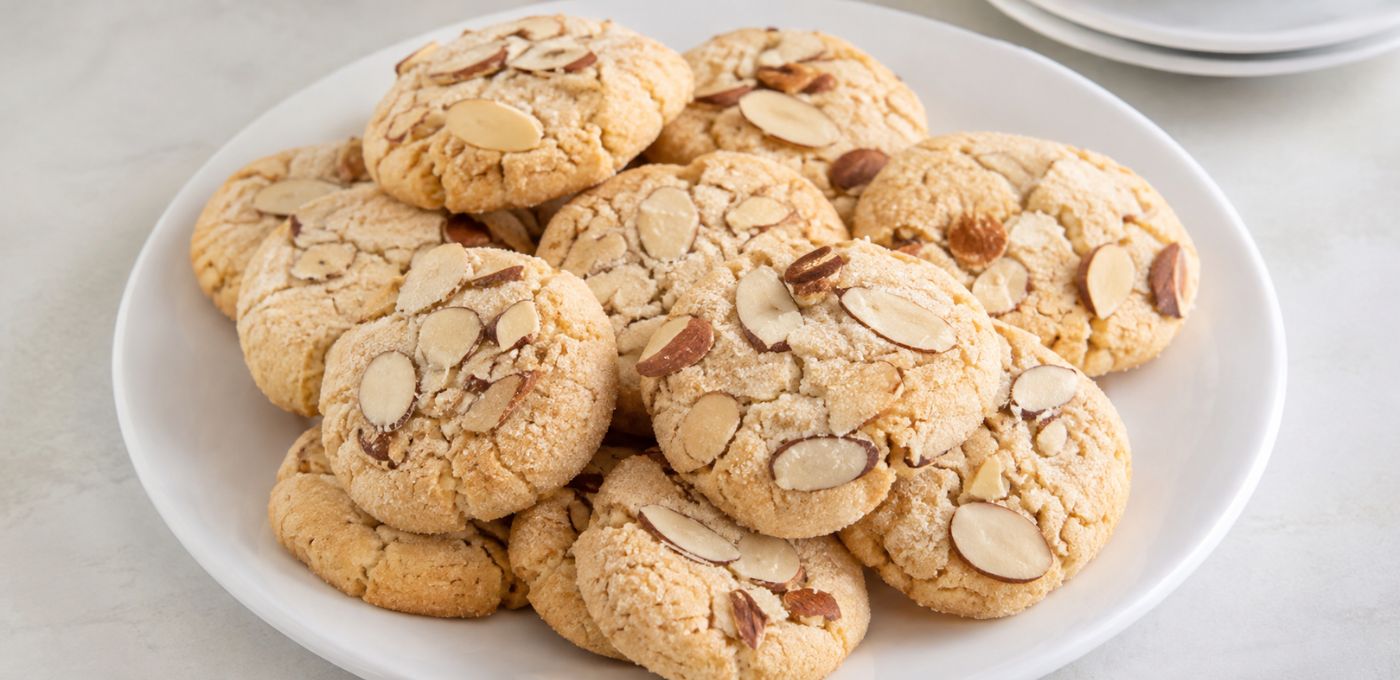 A pile of golden-brown almond cookies topped with sliced almonds, served on a white plate against a light marble background.
