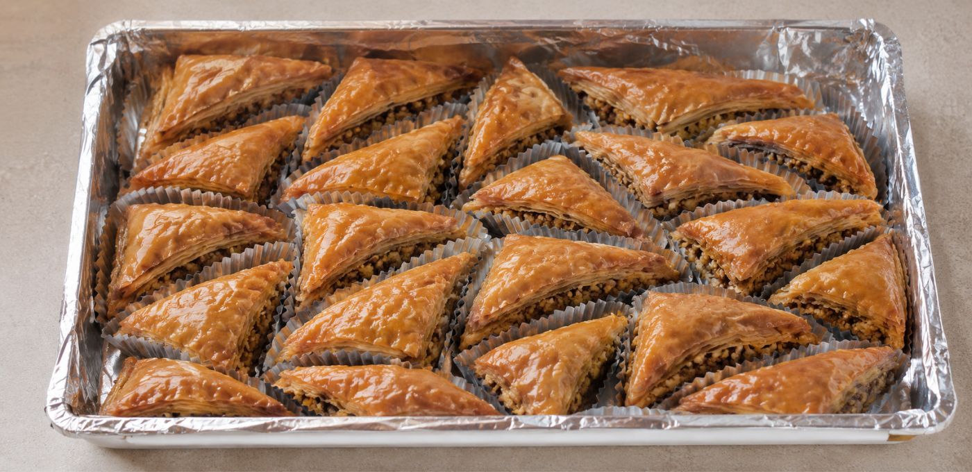 A tray of freshly baked, golden-brown Turkish baklava pastries arranged in a foil-lined baking pan on a neutral beige surface.