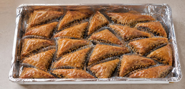 A tray of freshly baked, golden-brown Turkish baklava pastries arranged in a foil-lined baking pan on a neutral beige surface.