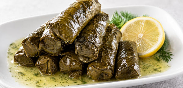 A pile of stuffed grape leaves (dolmas) served on a white plate with a lemon slice and dill garnish, set against a light gray background.