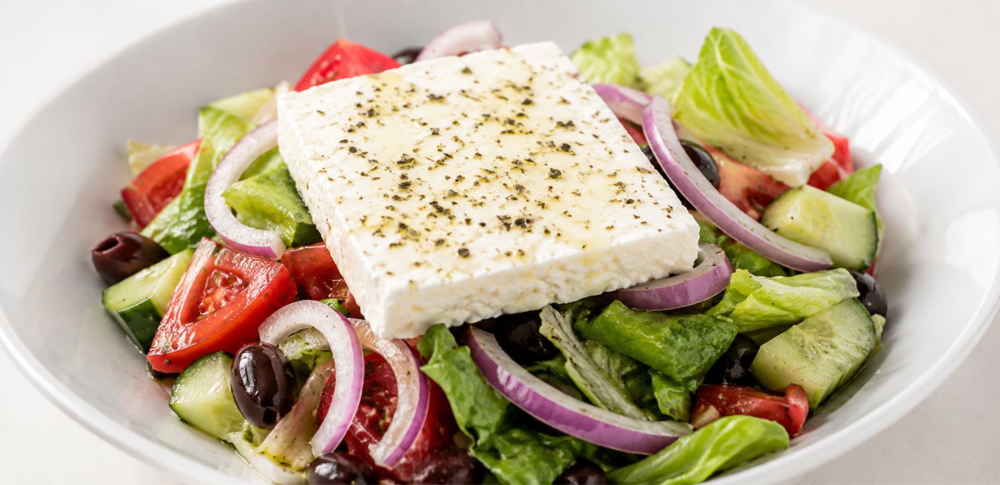 A close-up shot of a fresh Greek salad in a white bowl, featuring a large square of white feta cheese topped with herbs, surrounded by lettuce, cucumber, red onion, tomatoes, and black olives.