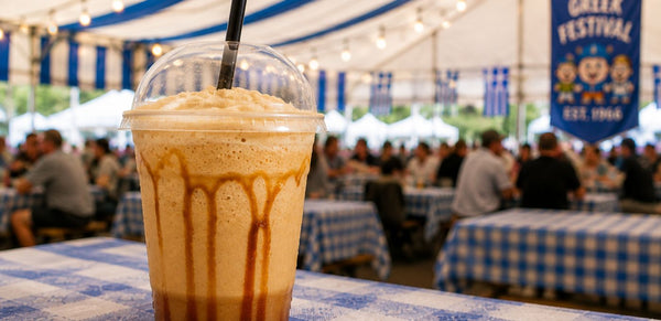 A frothy iced coffee or milkshake in a clear plastic cup with a black straw, topped with whipped cream and drizzled with caramel sauce, set on a blue-and-white checkered table at an outdoor festival under a tent.