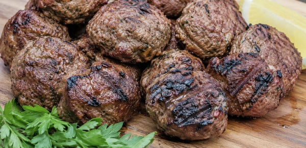 Grilled beef meatballs with charred marks, piled on a wooden board with parsley and a lemon wedge garnish.