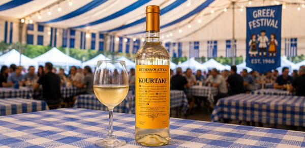 A bottle of Kourtaki white wine and a filled wine glass on a blue-and-white checkered tablecloth inside a large event tent, with a crowd and a 'Greek Festival' banner in the background.