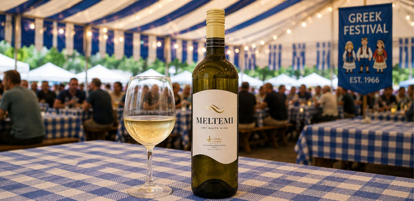 A bottle of Meltemi white wine and a filled wine glass on a blue-and-white checkered tablecloth at an outdoor Greek festival, with a blurred crowd and tents in the background.