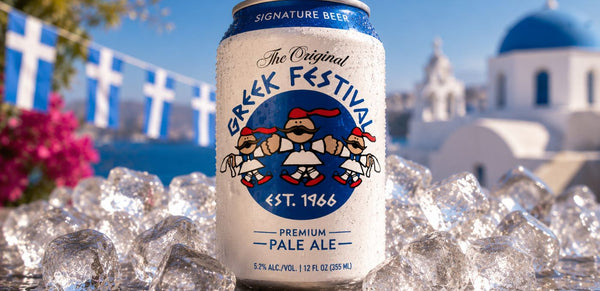 A can of Greek Festival Signature Beer in the center, surrounded by ice cubes, with a blurred background of a blue-domed white building and blue sky.