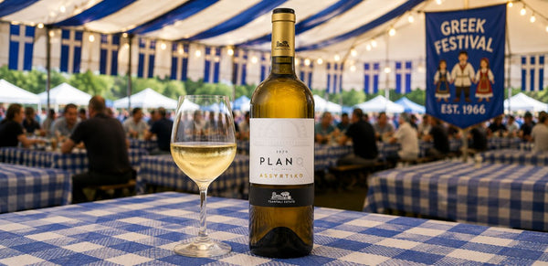 A bottle of Planos Alvyrtiko white wine and a filled wine glass on a blue-and-white checkered table at an outdoor Greek festival under a striped tent.