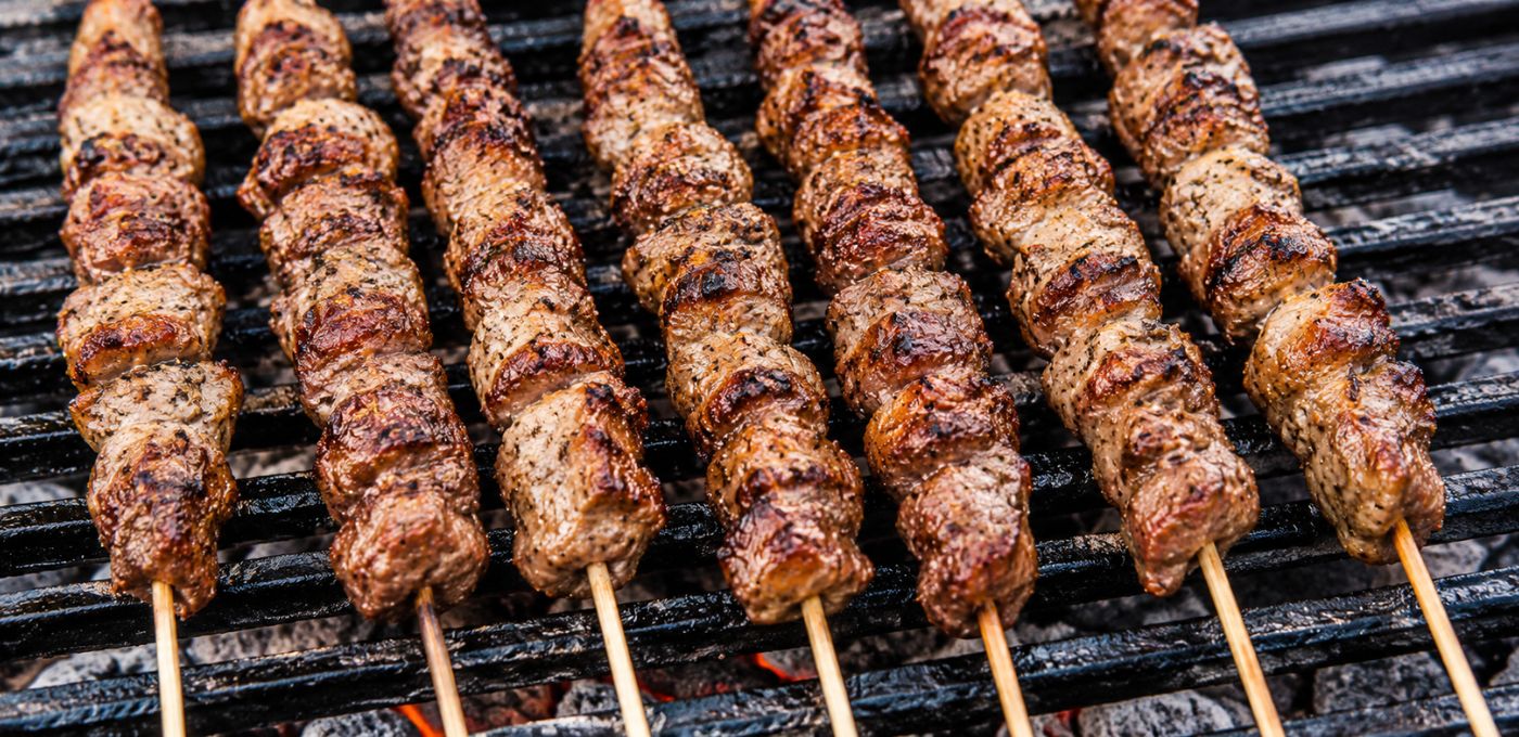 Grilled meat skewers with charred browned surfaces cooking on a black grill grate over coals.