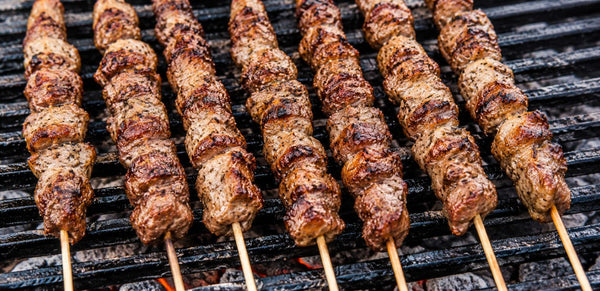 Grilled meat skewers with charred browned surfaces cooking on a black grill grate over coals.