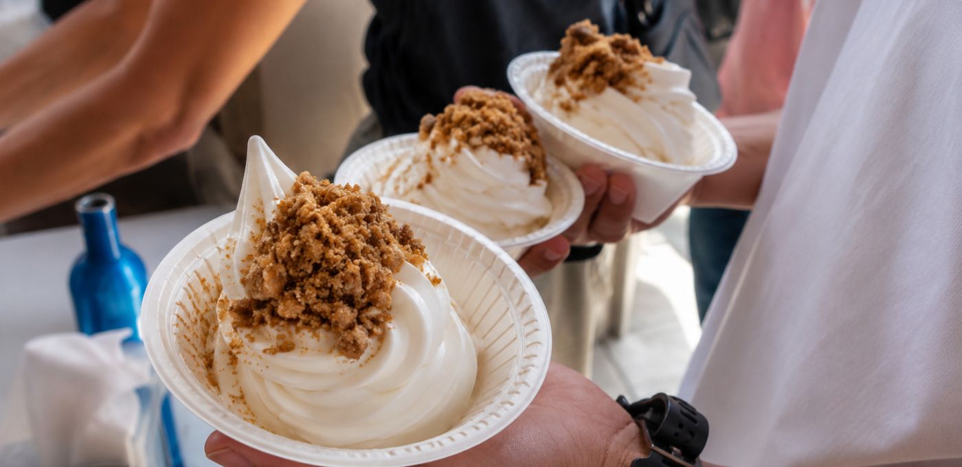 Three paper bowls of whipped cream or soft serve dessert topped with crumbly brown sugar, held by people at a dessert stand.
