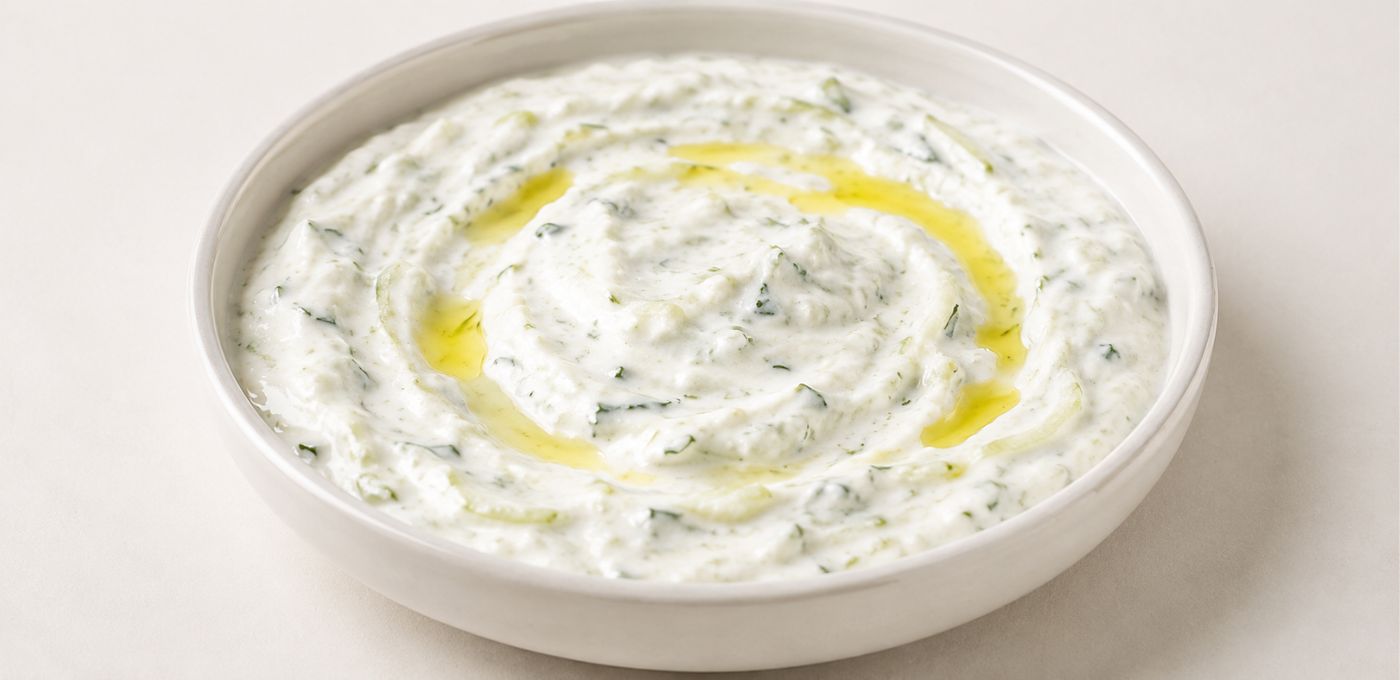 A white ceramic bowl filled with thick white tzatziki or yogurt dip, speckled with green herbs and drizzled with olive oil, on a light beige background.