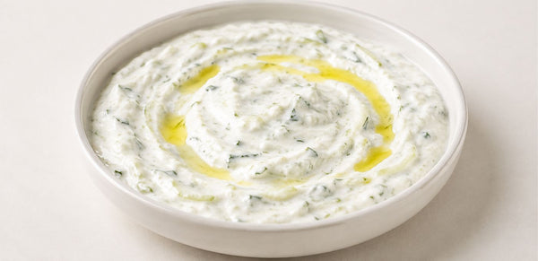 A white ceramic bowl filled with thick white tzatziki or yogurt dip, speckled with green herbs and drizzled with olive oil, on a light beige background.