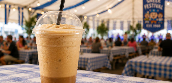 A frothy light brown iced coffee or smoothie in a clear plastic cup with a clear lid and black straw, sitting on a blue-and-white checkered table at an outdoor festival under a white tent.