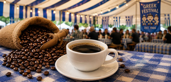 A white cup of black coffee on a saucer sits on a blue-and-white checkered tablecloth, with a burlap sack of coffee beans spilled beside it. In the background, a real event tent with people and a Greek Festival banner is visible.