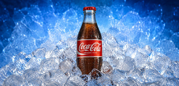 A classic glass bottle of Coca-Cola with a red cap and red label, standing upright in a bed of ice cubes against a blue icy background.