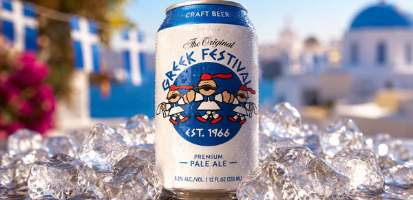A can of The Original Greek Festival Craft Beer, a pale ale, standing upright on a bed of ice cubes with a blurred Mediterranean background featuring white buildings and blue domes.