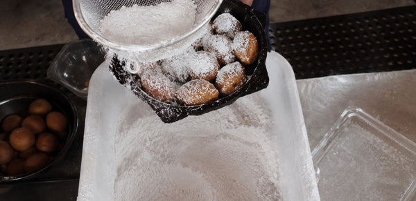 A close-up of freshly fried dough balls being dusted with white powdered sugar from a metal sieve, held over a large white tray filled with sugar in a kitchen setting.