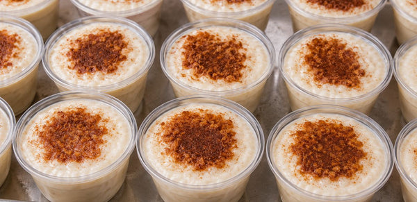Multiple small clear plastic cups filled with a creamy off-white dessert or pudding, each topped with a sprinkle of brown spice or cinnamon, arranged closely together.